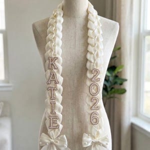 May include: Cream-coloured graduation sash with the name "KATIE" spelled out in sparkling letters and the year "2026" in pearl-like numbers. The sash is adorned with bows at the bottom. The sash is displayed on a mannequin.