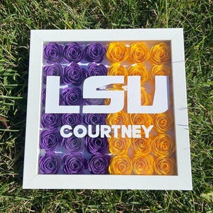 May include: A white frame with a purple and gold paper flower arrangement inside. The flowers are arranged in a grid pattern and spell out "LSU" with the name "COURTNEY" below.