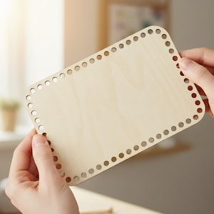 Puede incluir: Base rectangular de madera sin terminar para una cesta. La madera de color claro tiene esquinas redondeadas y una serie de agujeros espaciados uniformemente alrededor del perímetro. La base está sujeta por dos manos.