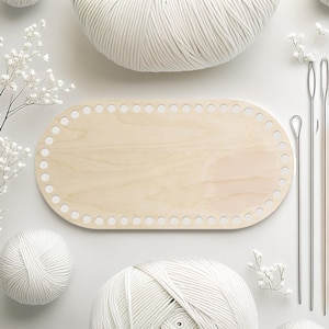 May include: A light-colored wooden oval base for a crochet bag, with evenly spaced holes around the edge. The image also shows white yarn balls, crochet hooks, and delicate white flowers, all arranged on a white surface.
