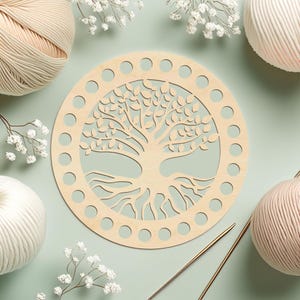 Può includere: Una base rotonda in legno chiaro con un design ad albero della vita, circondata da fori distanziati uniformemente. La base è circondata da gomitoli di filato nei toni del beige e del rosa, con rametti di fiori bianchi e ferri da maglia.