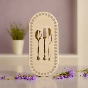 Può includere: Un telaio ovale in legno con un disegno di forchetta, coltello e cucchiaio. Il telaio è in legno chiaro e ha una serie di fori attorno al bordo.