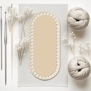 May include: A craft supplies flat lay featuring a beige oval wooden base with evenly spaced holes, two silver crochet hooks, two balls of cream-colored yarn, and sprigs of white flowers. The items are arranged on a white and gray background.