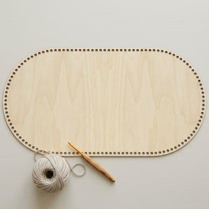 Op de afbeelding: Een licht houten ovale basis met gaten voor haakprojecten. Een bol grijs garen en een houten haaknaald zijn ook aanwezig, wat wijst op een knutselthema. De basis is ongeveer 30 cm lang.