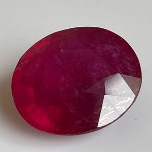 May include: A faceted, oval-shaped ruby gemstone. The gem is a deep, saturated red color with visible internal inclusions. The cut of the stone creates multiple reflective surfaces.