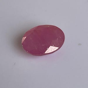Puede incluir: Gema de rubí ovalada y facetada. La gema es de color rojo rosado intenso y refleja la luz, mostrando su corte. La piedra se encuentra sobre un fondo blanco, resaltando su color y claridad. Ideal para la fabricación de joyas.