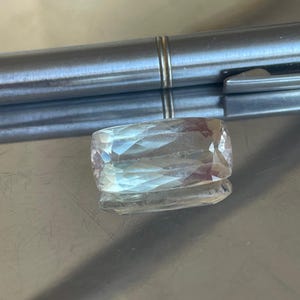 K&ouml;nnte beinhalten: Ein rechteckiger, facettierter Edelstein mit einer blassen, durchscheinenden Farbe. Der Stein liegt auf einer reflektierenden, metallischen Oberfl&auml;che, mit einem silbernen Stift im Hintergrund. Der Schliff des Steins erzeugt mehrere interne Reflexionen.