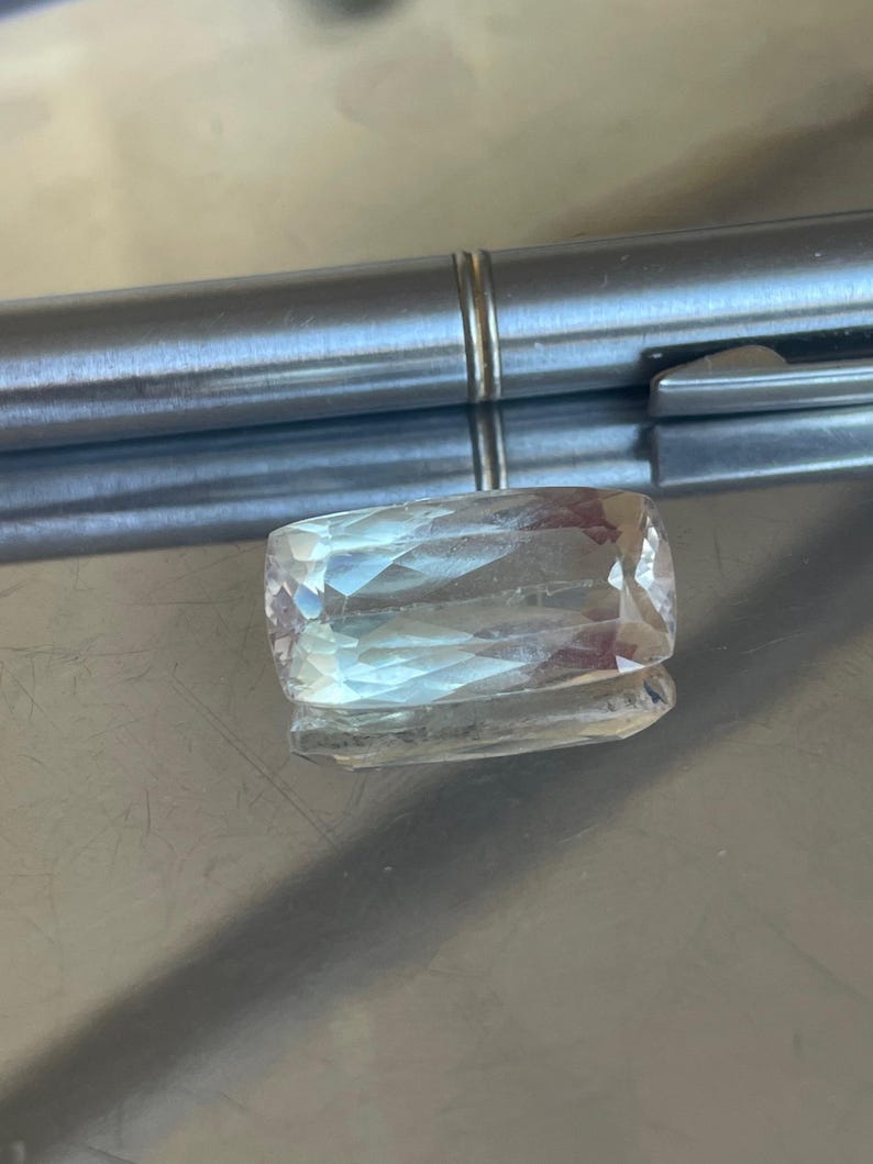 K&ouml;nnte beinhalten: Ein rechteckiger, facettierter Edelstein mit einer blassen, durchscheinenden Farbe. Der Stein liegt auf einer reflektierenden, metallischen Oberfl&auml;che, mit einem silbernen Stift im Hintergrund. Der Schliff des Steins erzeugt mehrere interne Reflexionen.