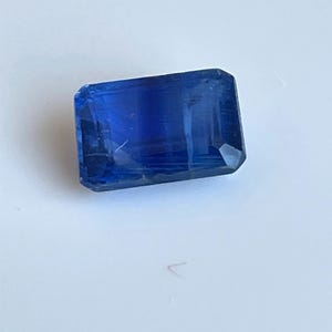 Könnte beinhalten: Ein rechteckiger, facettierter, tiefblauer Edelstein. Der Edelstein hat eine polierte Oberfläche mit sichtbaren inneren Strukturen und leichtem Glanz. Die Form ist ein längliches Rechteck mit geschnittenen Ecken, was darauf hindeutet, dass es sich um einen Saphir oder einen ähnlichen Edelstein handeln könnte.