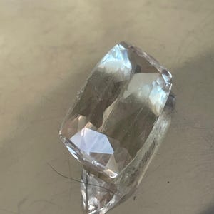 K&ouml;nnte beinhalten: Ein klarer, rechteckiger Edelstein mit mehreren Facetten, der Licht reflektiert. Der Edelstein hat einen leichten Gelbstich und liegt auf einer reflektierenden Oberfl&auml;che. Der Schliff des Edelsteins erzeugt einen funkelnden Effekt, geeignet f&uuml;r Schmuck.