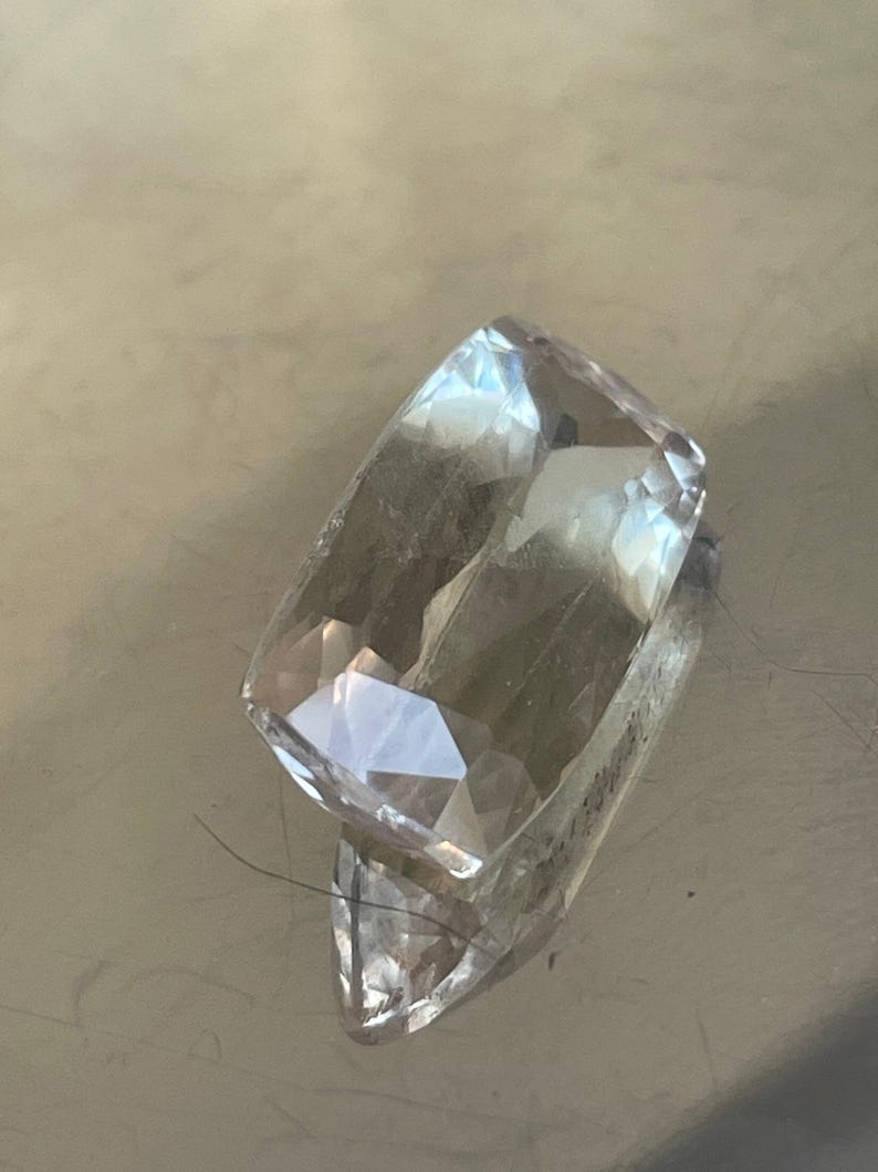 K&ouml;nnte beinhalten: Ein klarer, rechteckiger Edelstein mit mehreren Facetten, der Licht reflektiert. Der Edelstein hat einen leichten Gelbstich und liegt auf einer reflektierenden Oberfl&auml;che. Der Schliff des Edelsteins erzeugt einen funkelnden Effekt, geeignet f&uuml;r Schmuck.