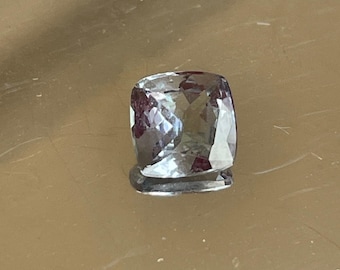 May include: A square-cut gemstone with a light blue and purple hue. The gem is faceted, reflecting light, and sits on a reflective surface. The gemstone is a jewelry component.