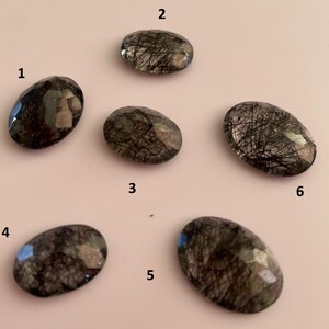 Pode incluir: Seis pedras preciosas ovais com corpos escuros e translúcidos e intrincadas inclusões pretas. As pedras são facetadas, captando a luz e exibindo uma gama de cores escuras. As pedras preciosas estão dispostas sobre uma superfície rosa claro.