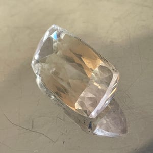 K&ouml;nnte beinhalten: Ein facettierter, rechteckiger Edelstein mit einem transparenten, hellbraunen Farbton. Der Edelstein hat mehrere Facetten, die das Licht reflektieren und einen funkelnden Effekt erzeugen. Der Stein liegt auf einer reflektierenden Oberfl&auml;che.