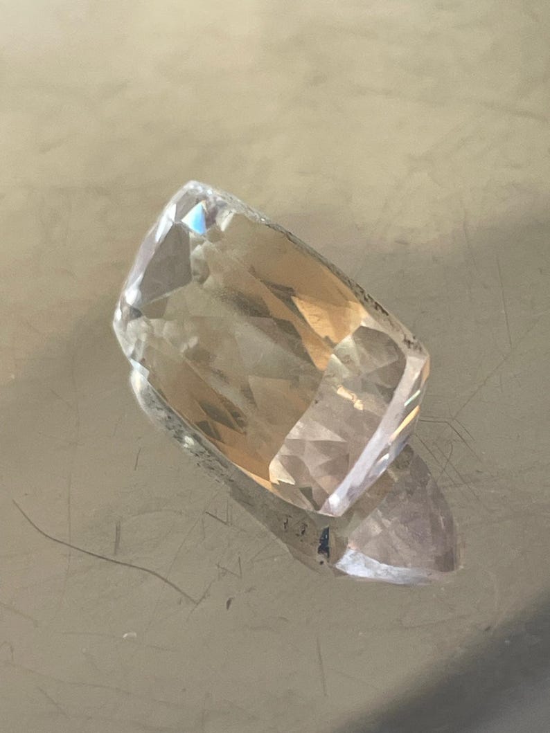 K&ouml;nnte beinhalten: Ein facettierter, rechteckiger Edelstein mit einem transparenten, hellbraunen Farbton. Der Edelstein hat mehrere Facetten, die das Licht reflektieren und einen funkelnden Effekt erzeugen. Der Stein liegt auf einer reflektierenden Oberfl&auml;che.