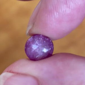 Può includere: Una piccola gemma rotonda, rosa-violacea, tenuta tra due dita. La gemma ha una superficie leggermente strutturata con punti riflettenti bianchi brillanti. Lo sfondo è di un colore marrone caldo sfocato.