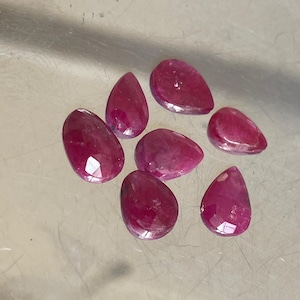 May include: Seven faceted rubies in teardrop and oval shapes. The gemstones are a deep red colour with a polished, reflective surface. The rubies are scattered on a light grey surface.