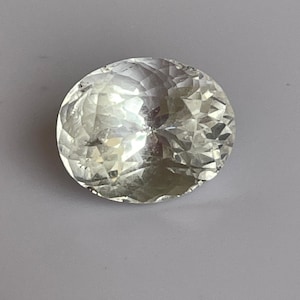 Può includere: Una gemma rotonda e sfaccettata con un aspetto chiaro e incolore. La gemma è tagliata per massimizzare la riflessione della luce, creando una brillantezza brillante. La pietra è posta su uno sfondo neutro.