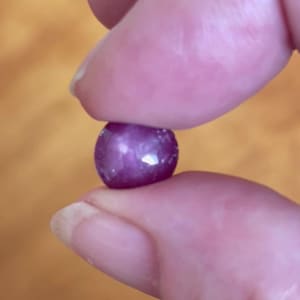 May include: A round, polished, deep pink gemstone, held between two fingers. The gem has a faceted surface that reflects light, creating bright highlights. The gem's colour is a rich, saturated pink, with a slight purplish hue.