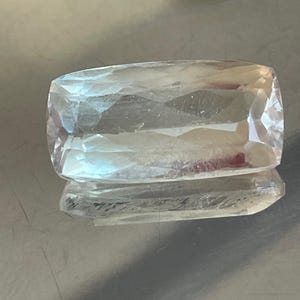 K&ouml;nnte beinhalten: Ein klarer, rechteckiger Edelstein mit facettierter Oberfl&auml;che. Der Edelstein ist farblos und reflektiert das Licht, wodurch ein funkelnder Effekt entsteht. Der Stein liegt auf einer reflektierenden Oberfl&auml;che und zeigt sein Spiegelbild.