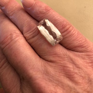 K&ouml;nnte beinhalten: Ein rechteckiger, facettierter Edelstein mit einem blassrosa Farbton. Der Edelstein wird in einer Hand gehalten, wodurch seine Klarheit und sein Schliff zur Geltung kommen. Die Facetten des Steins fangen das Licht ein und erzeugen einen funkelnden Effekt.