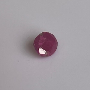 May include: A faceted, round, ruby gemstone. The gem is a deep pinkish-red color with multiple reflective surfaces. The gemstone is set against a plain white background.