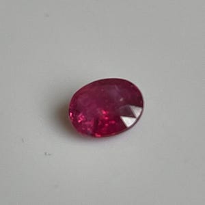 Könnte beinhalten: Ein oval geformter, facettierter Rubin-Edelstein. Der Rubin hat eine tiefe, satte rote Farbe mit polierter Oberfläche. Der Edelstein liegt vor einem weißen Hintergrund.