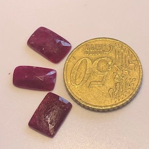 Peut inclure: Trois pierres précieuses rectangulaires, facettées, rouge foncé, à côté d'une pièce de 50 centimes d'euro. Les pierres précieuses ont une surface polie. La pièce est dorée et porte le chiffre 50 et le mot "CENT".