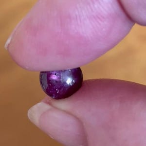 Op de afbeelding: Een kleine, ronde, dieppaarse edelsteen die tussen twee vingers wordt gehouden. De edelsteen heeft een gepolijst, reflecterend oppervlak, met hints van lichtere paarse en roze tinten. De belichting creëert highlights en schaduwen.