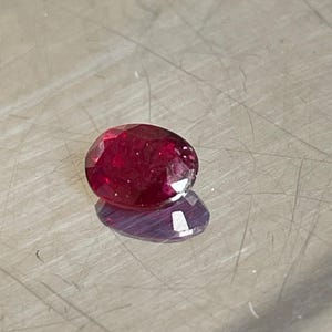 May include: A faceted, oval-shaped ruby gemstone. The gem is a deep, saturated red color with a reflective surface. The ruby is set against a light-colored, textured background.