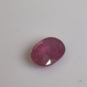 Może przedstawiać: Fasetowany, owalny kamień szlachetny rubinu. Klejnot ma głęboki, bogaty czerwony kolor z wypolerowaną powierzchnią. Fasetki wychwytują światło, tworząc efekt iskrzenia. Rubin jest osadzony na gładkim, jasnym tle.
