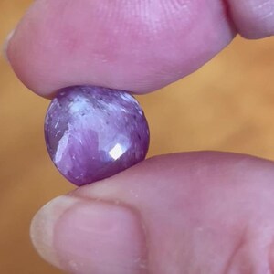 May include: A faceted, oval-shaped, purple gemstone held between two fingers. The gemstone has a polished surface with areas of lighter and darker purple hues. The background is a blurred, warm orange color.