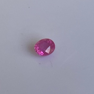 Może przedstawiać: Żywy, owalny różowy kamień szlachetny. Klejnot ma jasny, nasycony różowy kolor z fasetami, które odbijają światło. Kamień jest osadzony na gładkim, jasnym tle.