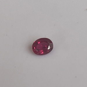 Peut inclure: Une pierre précieuse rubis de forme ovale. La gemme rouge foncé est posée sur un fond blanc. Les facettes de la pierre capturent la lumière, créant un effet scintillant. Ce rubis est un choix classique pour les bijoux.