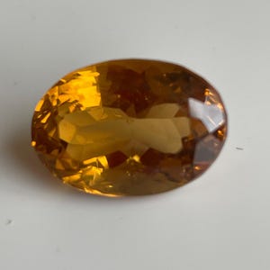 Può includere: Una pietra preziosa dal taglio ovale con una vibrante tonalità oro-arancio. La pietra presenta una superficie sfaccettata che riflette la luce, creando un brillante gioco di calde tonalità ambrate in tutta la sua struttura trasparente e lucida.