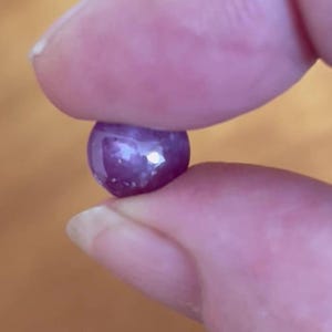 Könnte beinhalten: Ein kleiner, runder, dunkelvioletter Edelstein, der zwischen zwei Fingern gehalten wird. Der Edelstein hat eine glänzende Oberfläche mit reflektierenden Facetten, die das Licht einfangen. Der Hintergrund ist eine verschwommene, warmtonige Oberfläche.