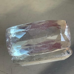 K&ouml;nnte beinhalten: Ein rechteckiger, facettierter Edelstein in blassblauer und klarer Farbe. Der Stein ist mit mehreren Facetten geschliffen, wodurch ein funkelnder Effekt entsteht. Der Edelstein wird auf einer reflektierenden Oberfl&auml;che pr&auml;sentiert, was seine Brillanz verst&auml;rkt. Geeignet f&uuml;r die Schmuckherstellung.
