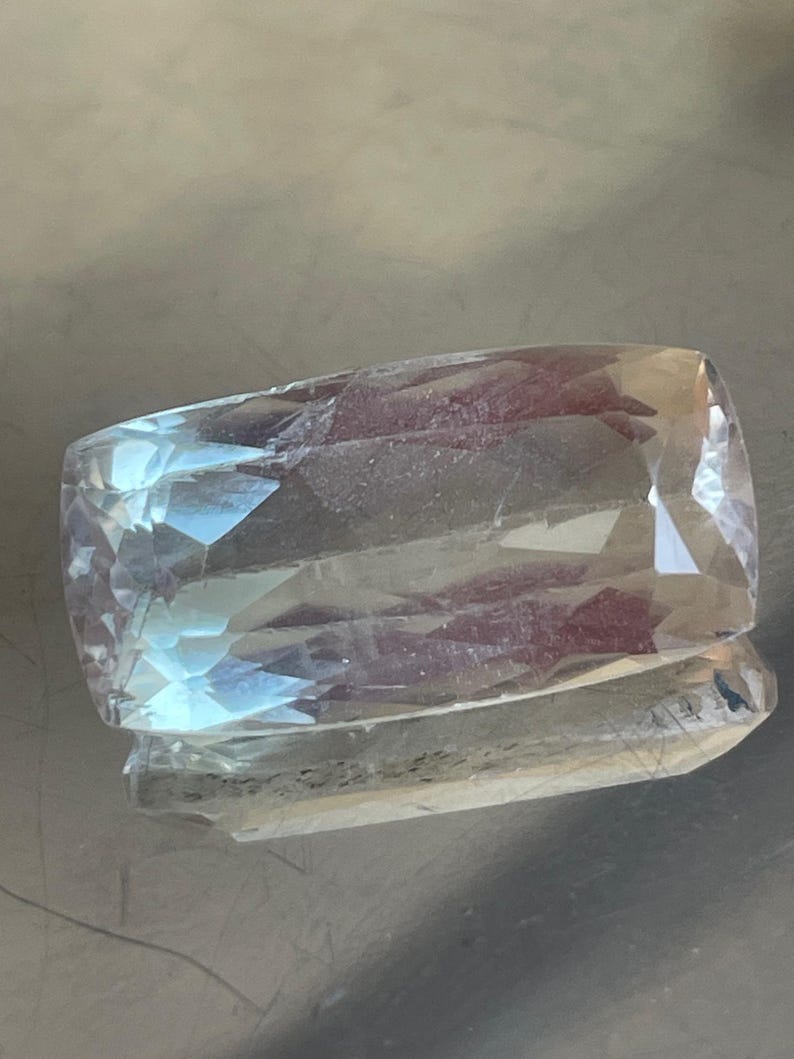 K&ouml;nnte beinhalten: Ein rechteckiger, facettierter Edelstein in blassblauer und klarer Farbe. Der Stein ist mit mehreren Facetten geschliffen, wodurch ein funkelnder Effekt entsteht. Der Edelstein wird auf einer reflektierenden Oberfl&auml;che pr&auml;sentiert, was seine Brillanz verst&auml;rkt. Geeignet f&uuml;r die Schmuckherstellung.
