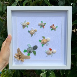 May include: A white framed picture with a white background featuring a sea-themed art piece made of sea glass and shells. The art piece depicts a group of sea turtles swimming in a cluster. The sea turtles are made of various shades of green, brown, and white sea glass and shells. The art piece is surrounded by a white mount and a white frame.