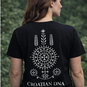 Könnte beinhalten: Schwarzes T-Shirt mit weißem Grafikdesign auf der Rückseite. Das Design zeigt ein kreisförmiges Muster mit einem Kreuz in der Mitte und anderen geometrischen Formen. Der Text "CROATIAN DNA" und "HERITAGE, NOT FASHION" ist darunter gedruckt.