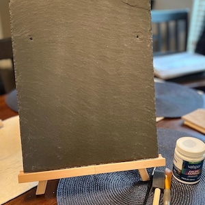 Puede incluir: Un caballete de pizarra con marco de madera, dos pinceles y un frasco de muestra de pintura blanca con la etiqueta "Valspar Color Sample".