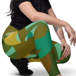 Może przedstawiać: Legginsy z kolorowym abstrakcyjnym wzorem w odcieniach zieleni, brązu i żółci. Legginsy są noszone z czarnym t-shirtem i czarnymi trampkami. Modelka kuca na białym tle.