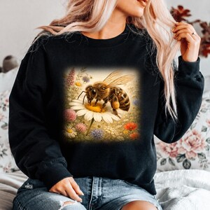 May include: A black long-sleeve sweatshirt featuring a central graphic print of a honeybee resting on a white daisy surrounded by various colorful wildflowers, including pink, red, and blue blossoms, set against a soft, light-colored background.