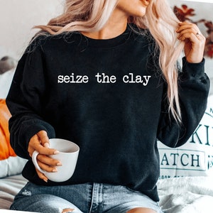 May include: A black sweatshirt with the text "seize the clay" printed in white on the front. The sweatshirt is being worn by a person holding a white mug.