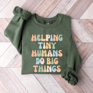 May include: An olive green sweatshirt with the text "HELPING TINY HUMANS DO BIG THINGS" in colorful, retro-style lettering. The long-sleeved top is made of a soft material and is suitable for casual wear.