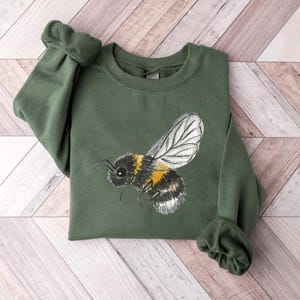 May include: A green sweatshirt featuring a detailed illustration of a bumblebee. The bee is depicted with black, yellow, and white markings, with a focus on the wings and body. The sweatshirt has a crew neck and long sleeves.