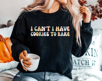 May include: A black sweatshirt with the text "I can't I have cookies to bake" in colorful, rounded letters.