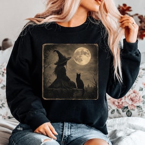 May include: A black long-sleeve sweatshirt featuring a square graphic print on the chest. The illustration depicts a silhouette of a person wearing a pointed hat sitting next to a cat under a large, bright full moon in a dark, wooded landscape.