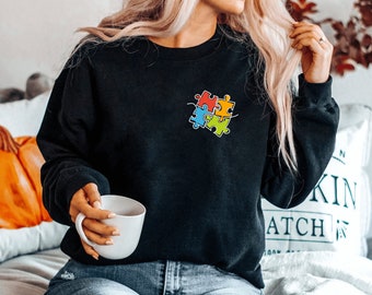 May include: A black sweatshirt with a colorful puzzle piece design on the front. The sweatshirt is being worn by a person with long blonde hair. The person is holding a white mug.