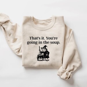 May include: A cream-colored sweatshirt with the text "That's it. You're going in the soup." and a black and white illustration of a frog in a witch's hat stirring a cauldron. The sweatshirt has long sleeves.